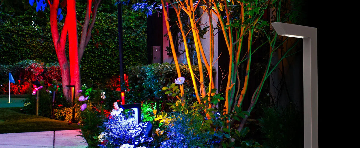 Modern outdoor landscape lighting at night, featuring a sleek white post-mounted LED light fixture on the right casting light upward, illuminating colorful garden plants, trees, and foliage in red, orange, blue, and purple hues.