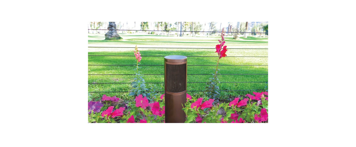 A bronze-colored outdoor bollard light fixture standing in a flowerbed with pink flowers, set against a blurred green park background.