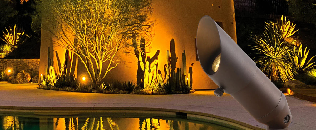 The beast an outdoor landscape light fixture with a wide angled head, shown in a nighttime poolside setting with warm yellow lighting illuminating cacti and trees against a stucco wall.