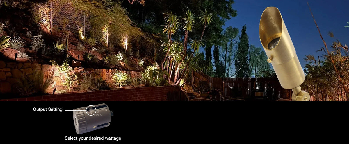 Outdoor landscape lighting fixture shown at night, with a close-up of the gold-toned spotlight on the right and a small inset showing an output setting control. The scene includes illuminated trees, plants, and stone walls against a dark blue evening sky.